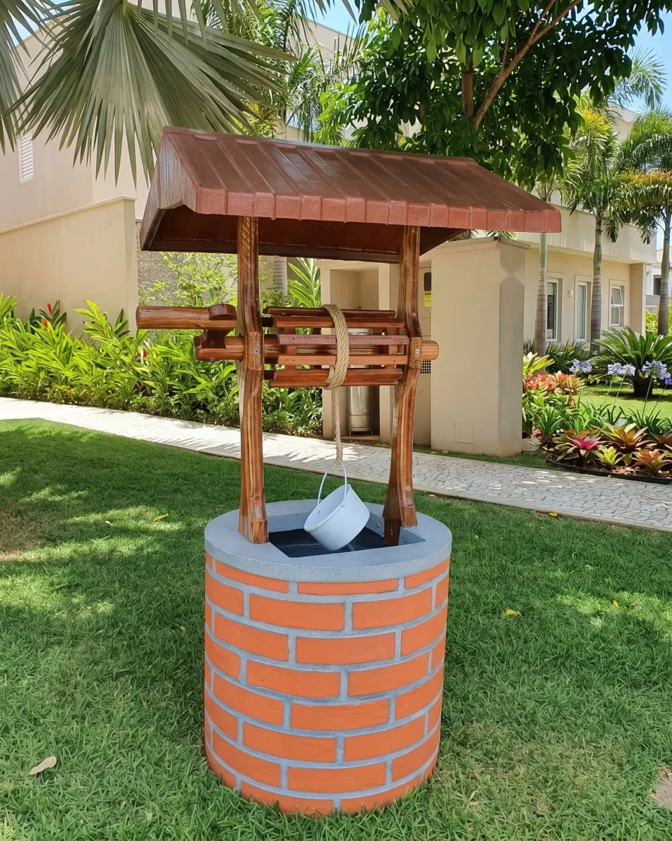 Fonte poço de jardim com base redonda de tijolinho laranja, colunas e sarilho de madeira escura, telhado de chapa e balde branco pendurado na corda, sobre grama